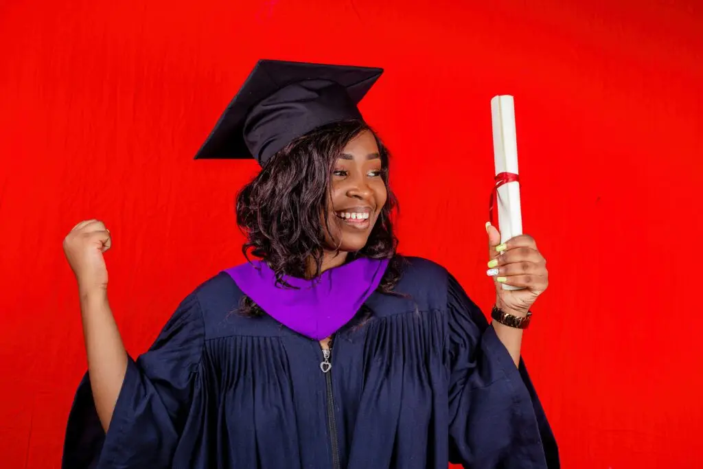 Smiling graduate in gown holding diploma against a vibrant red backdrop.
